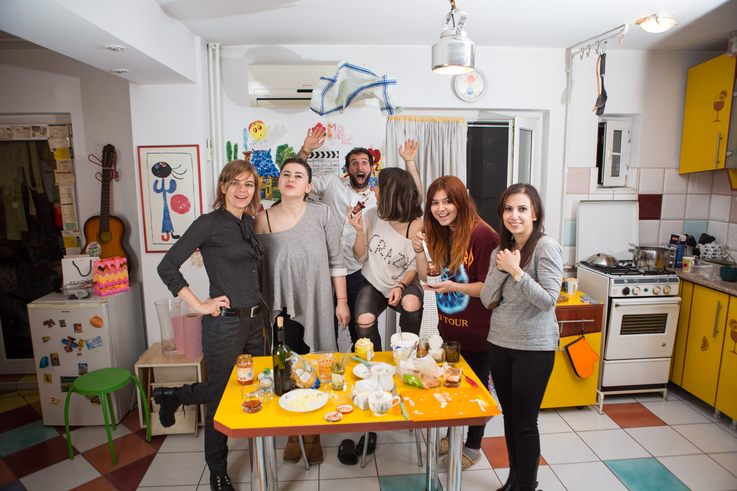 Invitații la cină pozând în picioare lângă masa portocalie, cu o chitară agățată pe peretele bucătăriei
