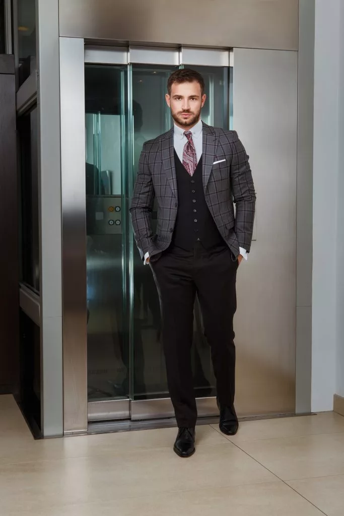 Bărbat în costum cu vestă, lângă ușile de lift — campanie de costume